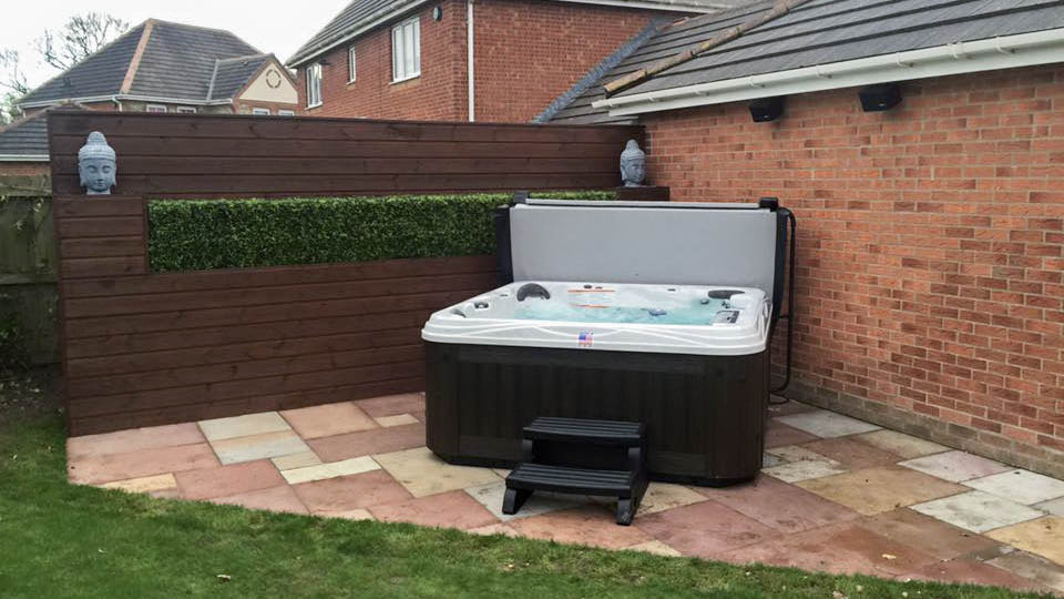 An Artesian spa next to a brick wall and a container garden hedge with a wooden slat exterior, buddha head statues on both ends of the container